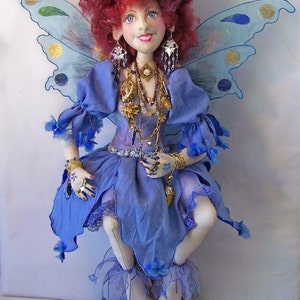 Pode incluir: Uma boneca de fada feita à mão com cabelo ruivo, um vestido azul e asas. Ela está usando joias de ouro e tem uma flor no cabelo.