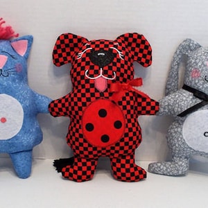 May include: Three handmade plush toys: a blue cat with pink ears and a white circle on its belly, a red and black checkered dog with a red circle on its belly, and a gray bunny with a black bow tie.
