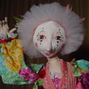May include: A handmade doll with a white face, red-tinged cheeks, and delicate facial features. The doll has a large, fluffy white headdress and is dressed in a colorful, patterned outfit with long sleeves. The doll's arm is raised, showcasing a detailed hand.