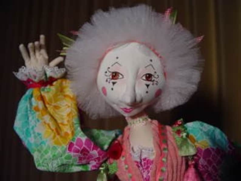 May include: A handmade doll with a white face, red-tinged cheeks, and delicate facial features. The doll has a large, fluffy white headdress and is dressed in a colorful, patterned outfit with long sleeves. The doll's arm is raised, showcasing a detailed hand.