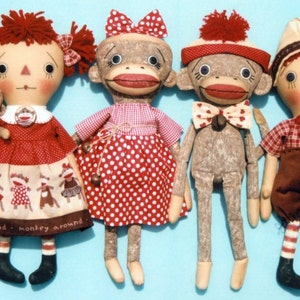 Puede incluir: Cinco muñecas hechas a mano, incluyendo una muñeca de trapo pelirroja, una muñeca mono con un vestido de lunares rojos y blancos, una muñeca mono con una corbata de lazo roja y blanca, y dos muñecas mono con camisas a cuadros rojos y blancos. Las muñecas están hechas de tela y tienen un aspecto vintage.