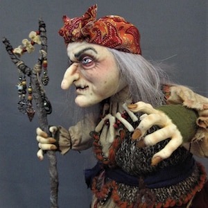 May include: A handmade doll with a long white wig, a red and orange paisley bandana, and a long, gnarled stick. The doll is wearing a brown and white striped shirt, a brown and gray vest, a blue sash, and a red and orange skirt. The doll has a long, crooked nose and a fierce expression.