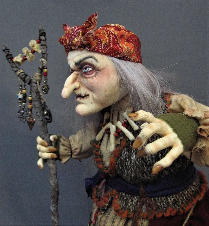 May include: A handmade doll with a long white wig, a red and orange paisley bandana, and a long, gnarled stick. The doll is wearing a brown and white striped shirt, a brown and gray vest, a blue sash, and a red and orange skirt. The doll has a long, crooked nose and a fierce expression.