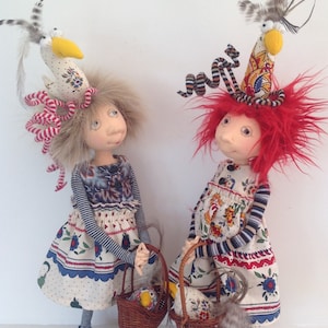 May include: Two handmade dolls with red and blonde hair wearing colorful dresses and hats. Each doll is holding a basket with a bird inside.