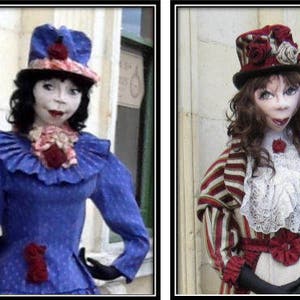 May include: Two dolls wearing vintage-style clothing and hats. The doll on the left is wearing a blue dress with a white collar and a blue hat with a red flower. The doll on the right is wearing a red and white striped dress with a white lace collar and a red and white hat with a red flower.