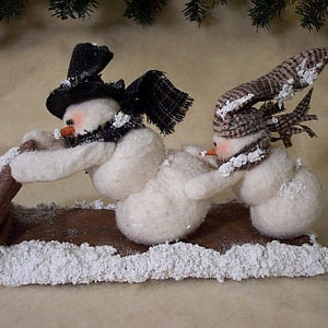 May include: Two snowmen made of white felt are riding on a brown sled. The snowman in the front is wearing a black top hat and a black scarf. The snowman in the back is wearing a brown and white patterned hat. The sled is covered in white faux snow.