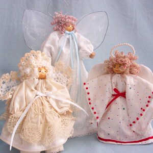 May include: Three handmade fabric angels with white dresses and wings. The angel on the left has a lace dress and white wings. The angel in the center has a white dress with a blue ribbon and sheer wings. The angel on the right has a white dress with red polka dots and red ribbon, and white wings.