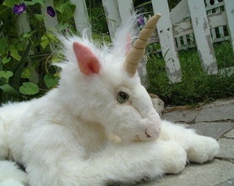 Könnte beinhalten: Ein weißes Plüsch-Einhorn mit einem langen, cremefarbenen Horn. Das Einhorn liegt auf der Seite, der Kopf ruht auf den Vorderpfoten. Das Einhorn hat ein rosa Ohr und grüne Augen.