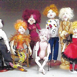 May include: A group of nine dolls with different hairstyles, clothing, and accessories. The dolls are made of fabric and have painted faces. They are all standing or sitting on a white background.