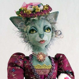 Może przedstawiać: Fantazyjna lalka z niebiesko-zieloną twarzą i szarym futrem, ubrana w różową i fioletową sukienkę z haftowanymi kwiatami i koroną z ptaków i kwiatów.