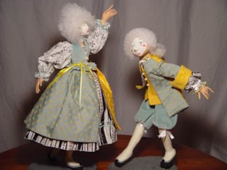 May include: Two handmade dolls dressed in 18th-century attire. The female doll wears a light blue dress with a yellow sash and a white wig. The male doll is in a matching outfit with a yellow jacket and a white wig. Both dolls have painted faces.