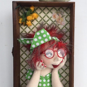 Op de afbeelding: Een stoffen pop met rood haar, een rode bril en een groen outfit met witte stippen bevindt zich in een houten doos. De pop rust met de kin op de handen. Boven de pop zitten twee groene keramische vogels op de rand van de doos met gele bloemen.