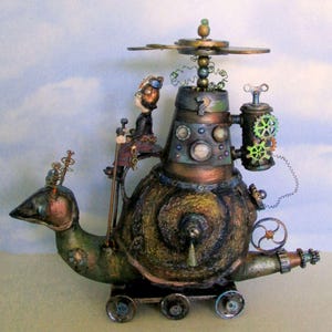 Op de afbeelding: Een grillig steampunk-slakkenbeeld met een miniatuurfiguur dat er bovenop zit. De slak heeft een koperkleurige schelp met ingewikkelde details en een propeller bovenop. De figuur draagt een bruin jasje en een hoed.