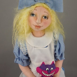May include: A handmade doll dressed as Alice in Wonderland, with blonde curly hair, a blue and white gingham dress, and a white apron. She is holding a purple and pink plush cat with a wide grin.