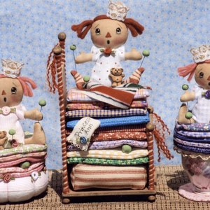 May include: Three handmade rag dolls dressed in white dresses with pink accents, each sitting on a stack of colourful fabric. The dolls are wearing crowns and have red hair. The dolls are sitting on a bed, a stack of fabric, and a teacup.
