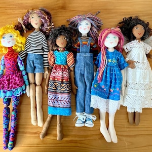 May include: Six handmade fabric dolls arranged in a row. The dolls feature diverse yarn hair colors, including yellow, purple, black, multicolored, pink, and dark brown, and wear various outfits like patterned dresses, denim overalls, and lace.
