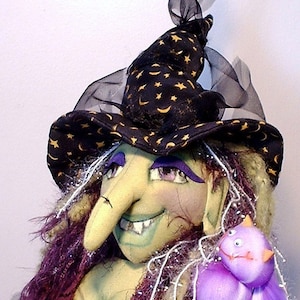 May include: A handmade Halloween witch doll with a green face, purple hair, and a black hat adorned with gold stars and moons. The witch has a long nose, a toothy grin, and is holding a purple creature. The doll is a festive decoration.