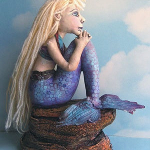 Peut inclure: Une figurine de sirène faite à la main avec de longs cheveux blonds, une queue violette et bleue et un soutien-gorge en coquillage. La sirène est assise sur un rocher brun texturé.