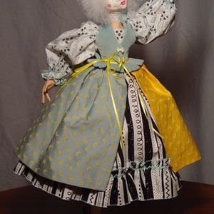 May include: A handmade doll with a white wig and a pale face. The doll wears a light blue and yellow dress with a black and white patterned skirt. The doll's sleeves are white with a black floral pattern. The doll is standing on a gray base.