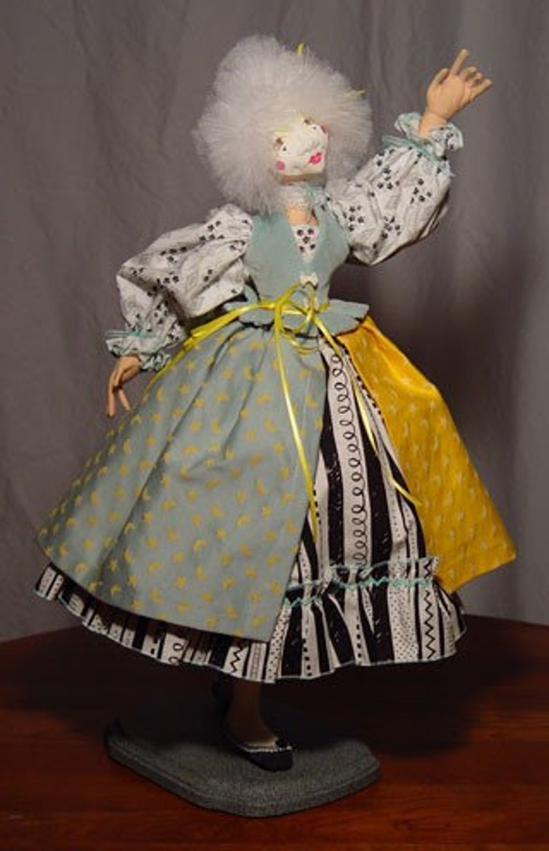May include: A handmade doll with a white wig and a pale face. The doll wears a light blue and yellow dress with a black and white patterned skirt. The doll's sleeves are white with a black floral pattern. The doll is standing on a gray base.