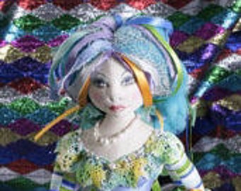Puede incluir: Una muñeca artesanal con una expresión serena, adornada con un tocado azul y verde y un atuendo a juego. La muñeca presenta un collar de perlas y detalles intrincados, mostrando un diseño caprichoso y artístico.