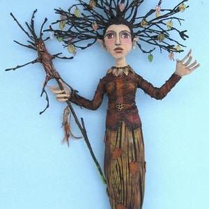 Pode incluir: Uma escultura artesanal e fantasiosa de um espírito da floresta com uma coroa de galhos de árvores e um bastão de madeira. A figura tem um rosto detalhado, roupas marrons e uma saia com detalhes de folhas. A escultura está em um fundo azul claro.