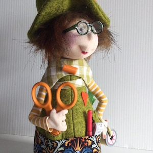 Può includere: Una bambola fatta a mano che raffigura una sarta, con un cappello verde con spilli, occhiali e forbici arancioni. La bambola ha una gonna fantasia e maniche a righe, con un metro a nastro e altri strumenti di cucito.