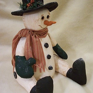 May include: A white snowman with a black top hat, green scarf, and black mittens. The snowman has a carrot nose and black eyes. The snowman is sitting on a white surface.