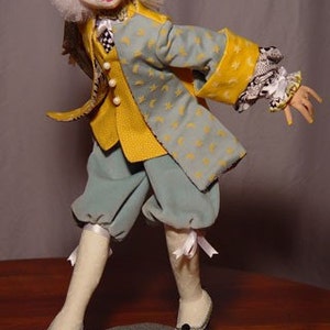 May include: A handmade doll dressed in a yellow and blue jacket with star and crescent moon patterns. The doll has a white wig, rosy cheeks, and is wearing blue pants, white stockings, and black shoes. The doll is standing on a gray base.