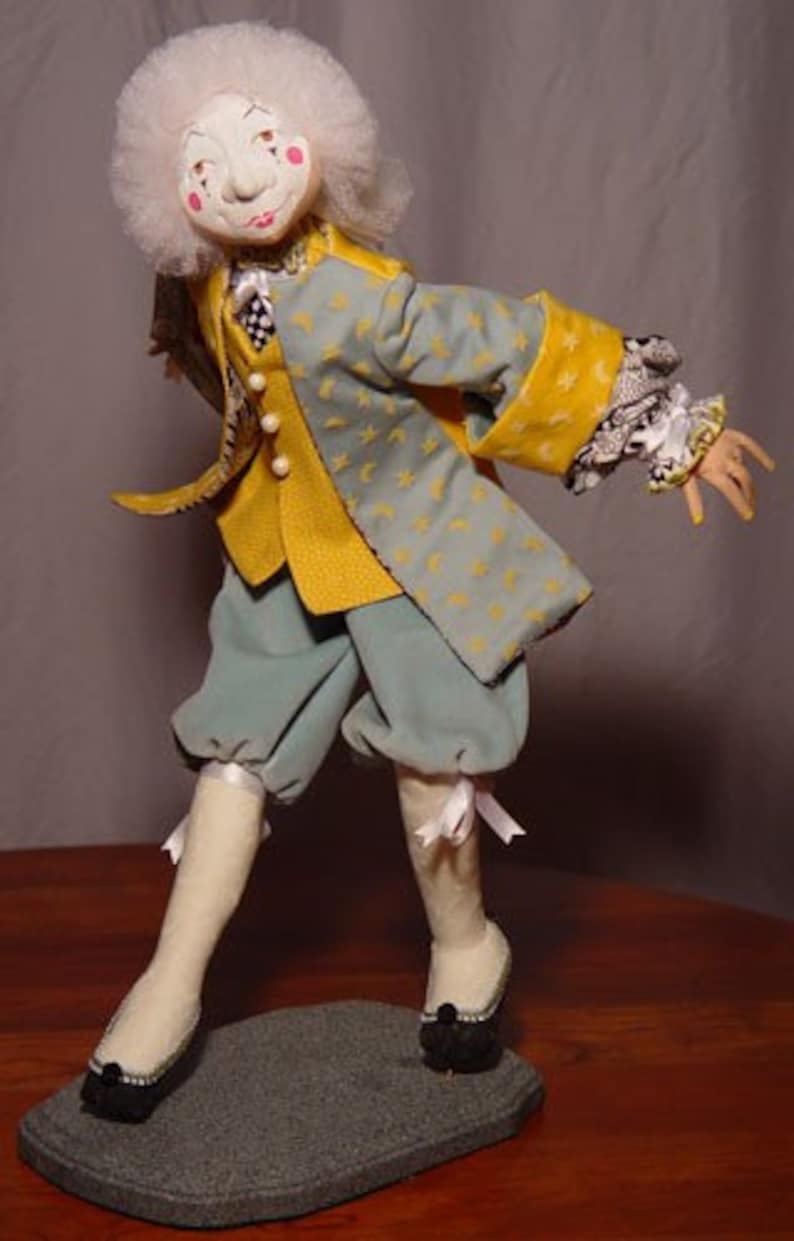 May include: A handmade doll dressed in a yellow and blue jacket with star and crescent moon patterns. The doll has a white wig, rosy cheeks, and is wearing blue pants, white stockings, and black shoes. The doll is standing on a gray base.