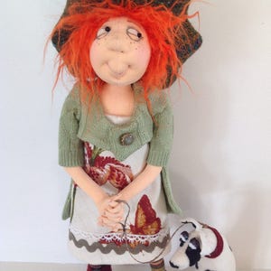 May include: A handmade doll with red hair, wearing a green cardigan, a floral dress, and a brown and green hat. The doll is holding a leash attached to a small black and white dog.