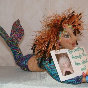 May include: A mermaid doll with long, curly, orange hair, wearing a blue and green patterned dress and a colorful tail. She is holding a card that says "I'm creating through life, how about you?"