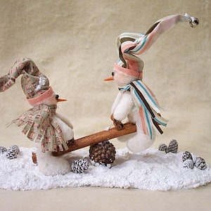 Könnte beinhalten: Zwei Plüsch-Schneemannfiguren auf einer Wippe, eine trägt einen rosa-weiß gestreiften Hut und die andere einen braunen und weißen karierten Hut. Die Schneemänner stehen auf einer weißen schneebedeckten Oberfläche mit Tannenzapfen.