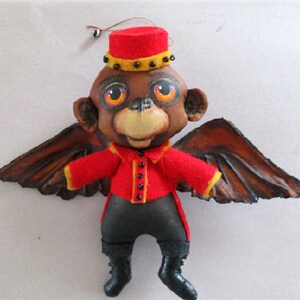 May include: A plush monkey doll wearing a red and yellow uniform with black buttons and brown wings. The monkey has brown fur, black eyes, and a black nose.