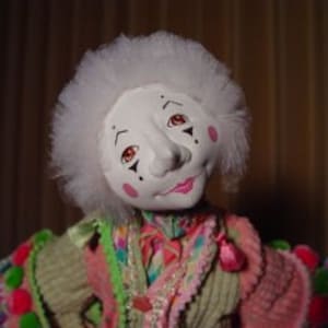 May include: A handmade clown doll with a white face, rosy cheeks, and a pink mouth. The doll has a white, fluffy wig and is wearing a colorful, patchwork-style outfit with pink, green, and orange accents. The doll's eyes are brown.