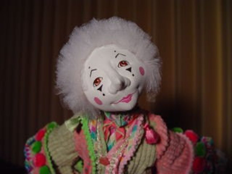 May include: A handmade clown doll with a white face, rosy cheeks, and a pink mouth. The doll has a white, fluffy wig and is wearing a colorful, patchwork-style outfit with pink, green, and orange accents. The doll's eyes are brown.