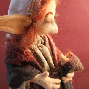 May include: A handmade felt doll with a whimsical design. The doll features a light-colored hat with a braided trim, long pointed ears, and red hair. It wears a layered outfit in shades of red, gray, and beige, and holds a smaller doll.