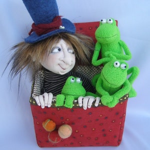 May include: A whimsical doll with brown hair wearing a blue top hat sits in a red box with three green frog dolls. The box has a polka dot pattern.