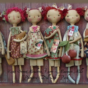 Peut inclure: Six poupées de chiffon faites à la main avec des cheveux en laine rouge, chacune tenant un fruit ou un légume différent. Les poupées sont habillées de tissus d'inspiration vintage avec des motifs floraux et géométriques. Les poupées sont alignées sur un fond en bois. Le texte "Sweet Meadows Farm -  Maureen Mills Primitive Doll Set - 6 Pack" est visible en bas de l'image.
