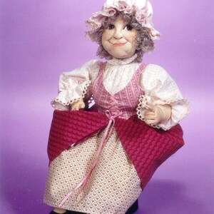 May include: A handmade doll with gray hair, wearing a pink and white dress with a red and white patterned skirt. The doll is sitting on a purple background.