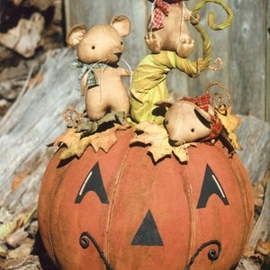 May include: Three brown fabric mice are perched on a large orange pumpkin with a black painted jack-o'-lantern face. The pumpkin is sitting on a brown log.