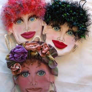 May include: Three fabric face-shaped purses with blue eyes and red lips. One has red, fluffy hair, another has black and green hair, and the third has a floral headdress with dangling beaded earrings. The purses are on a white background.