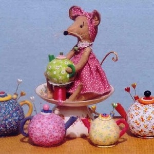 May include: A pink fabric mouse doll holding a green and white teapot pincushion. The mouse is sitting on a white pedestal with four other pincushions shaped like teapots. The teapots are decorated with floral patterns in blue, pink, yellow, and white.