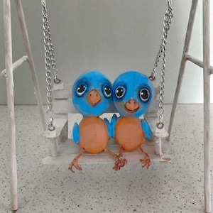 May include: Two blue bird figurines with large eyes and orange beaks are sitting on a white swing set. The swing set is made of metal and has a white wooden seat.