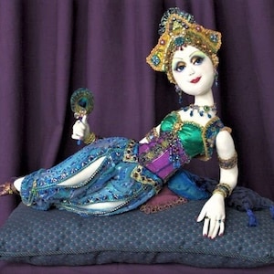 May include: A handmade doll with a jeweled headdress, lying on a blue and green patterned cushion. The doll is wearing a blue and green outfit with gold accents and is holding a peacock feather fan.