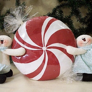Könnte beinhalten: Zwei weiße Schneemannfiguren mit blau-weiß karierten Schals und Kleidern halten einen großen roten und weißen Pfefferminzbonbon. Der Bonbon ist in durchsichtige Cellophanfolie verpackt.
