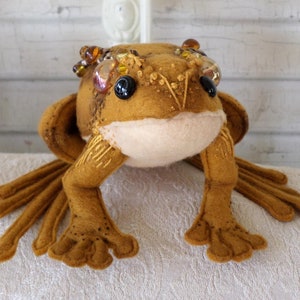 May include: A brown felt frog with beaded eyes and a crown of gold and amber beads. The frog has long, slender legs and a wide, flat body.