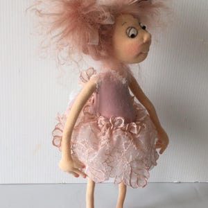 May include: A handmade doll with a whimsical design. The doll features a pink dress with a lace overlay, matching pink shoes, and fluffy pink hair. The doll has a light skin tone and a slightly surprised expression.