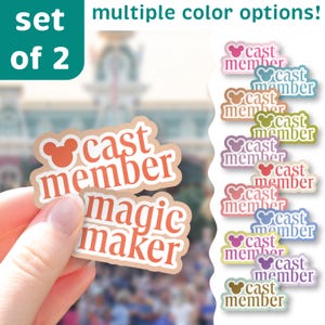 Puede incluir: Un juego de dos pegatinas con el texto "cast member magic maker" en naranja y blanco sobre un fondo beige. Las pegatinas tienen un icono de la cabeza de Mickey Mouse. La imagen también muestra múltiples opciones de color de la pegatina "cast member".