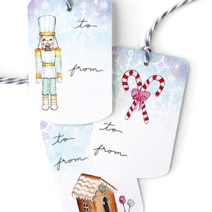 May include: Three watercolor Christmas gift tags with blue and white backgrounds. The first tag features a nutcracker, the second tag features candy canes, and the third tag features a gingerbread house. Each tag has the words "to" and "from" written on them.